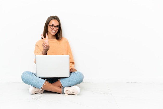 Jeune femme avec un ordinateur portable assis sur le sol souriant et montrant le signe de la victoire