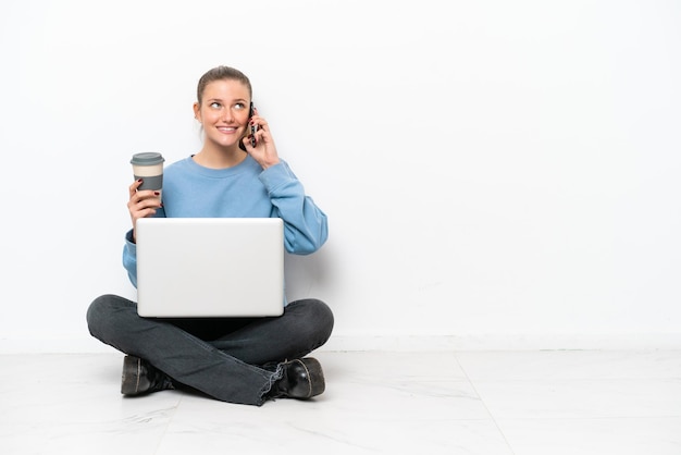 Jeune femme avec un ordinateur portable assis sur le sol tenant du café à emporter et un mobile