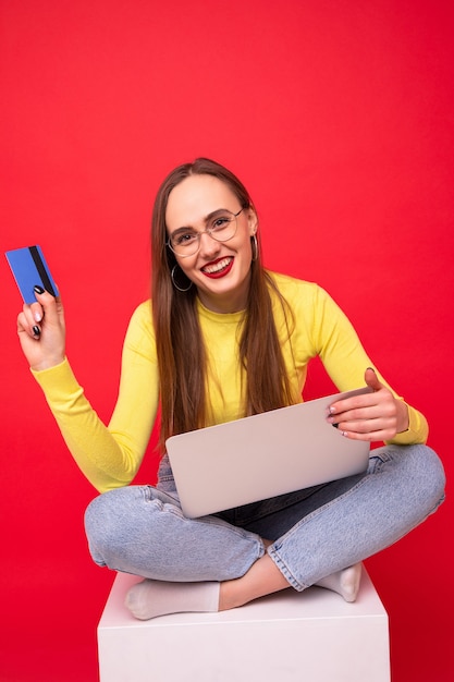 Jeune femme avec ordinateur portable et carte bancaire sur fond rouge