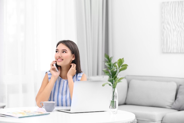 Jeune femme avec un ordinateur portable moderne parlant par téléphone assis à table dans la chambre