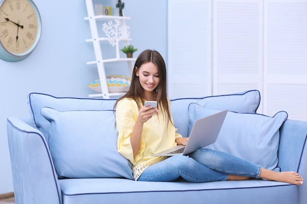 Jeune femme avec ordinateur portable moderne et téléphone assis sur un canapé à la maison