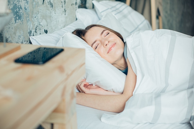 Jeune femme paisible allongée les yeux fermés dans son lit et souriant joyeusement