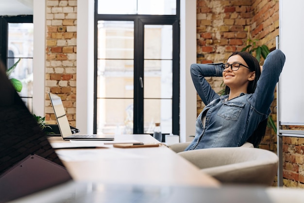 Photo jeune femme paisible se sentant détendue au bureau avec les yeux fermés et se tenant la main derrière la tête à l'intérieur du bureau