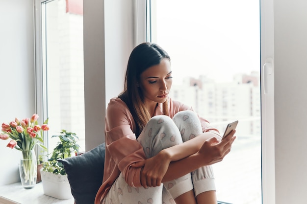 Jeune femme paresseuse en pyjama confortable utilisant un téléphone intelligent tout en se reposant sur le rebord de la fenêtre à la maison