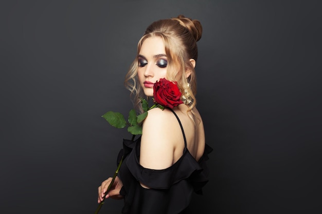 Une jeune femme parfaite avec des cheveux blonds et du maquillage tenant une fleur de rose rouge sur un fond noir