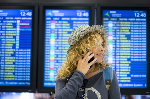 Jeune femme parlant au téléphone portable à l'aéroport lors d'un voyage. Passager féminin au chapeau regardant loin et parlant au téléphone portable devant l'affichage numérique à l'aéroport