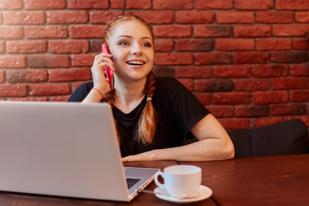 Jeune femme parlant au téléphone tout en travaillant en ligne via un ordinateur portable au café