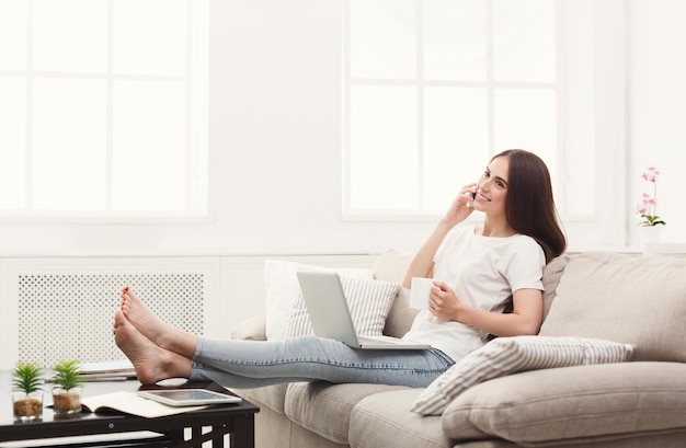 Jeune femme parlant sur mobile, vérifiant les médias sociaux et buvant du café à la maison sur le canapé. Fille aux cheveux noirs dans un espace de copie décontracté