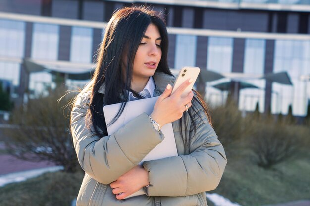 Une jeune femme parle au téléphone et tient un ordinateur portable à l'extérieur