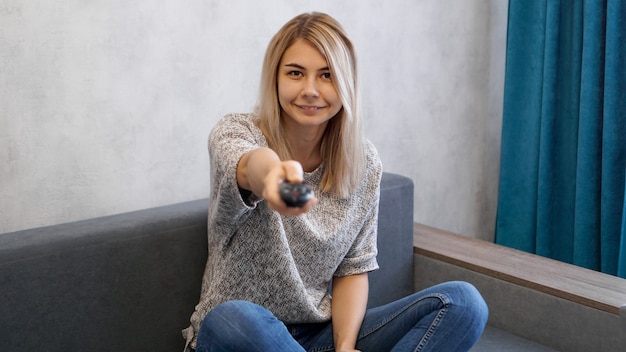 Jeune femme passe les chaînes de télévision avec la télécommande. Elle sourit et regarde la caméra