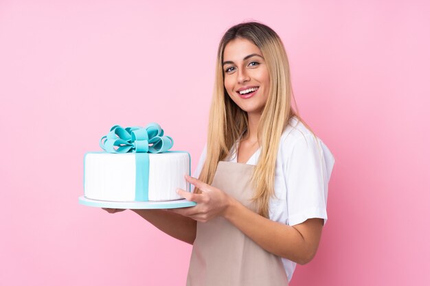 Jeune femme pâtissière avec un gros gâteau sur un mur rose isolé souriant beaucoup