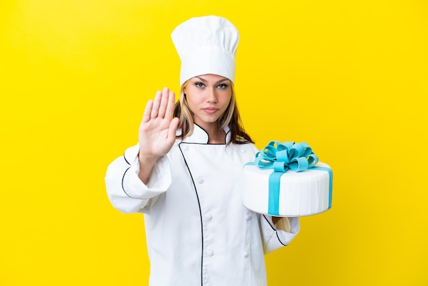 Jeune femme pâtissière russe avec un gros gâteau isolé sur fond jaune faisant un geste d'arrêt