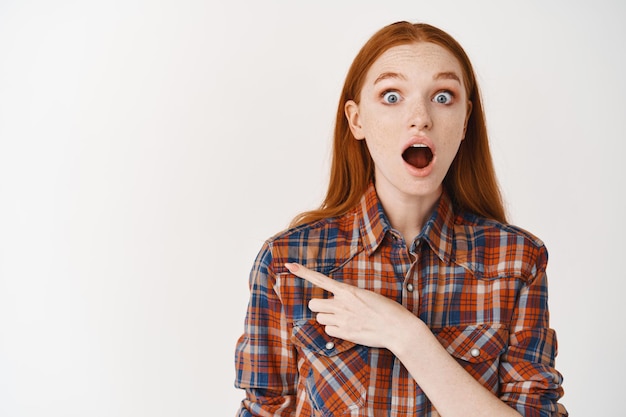 Jeune femme à la peau pâle et aux cheveux roux, l'air surpris à l'avant, pointant le doigt vers la gauche sur une offre promotionnelle incroyable, debout sur un mur blanc