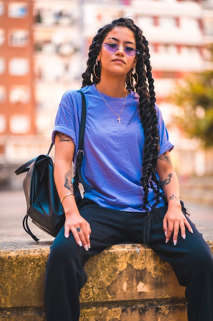 Jeune femme à la peau sombre avec de longues tresses et des lunettes violettes, assise sur un banc dans la ville avec son sac à dos en bandoulière