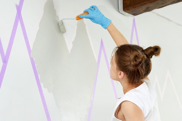 Jeune femme peignant un mur avec un rouleau à peinture et utilisant du ruban adhésif en se tenant debout sur une échelle