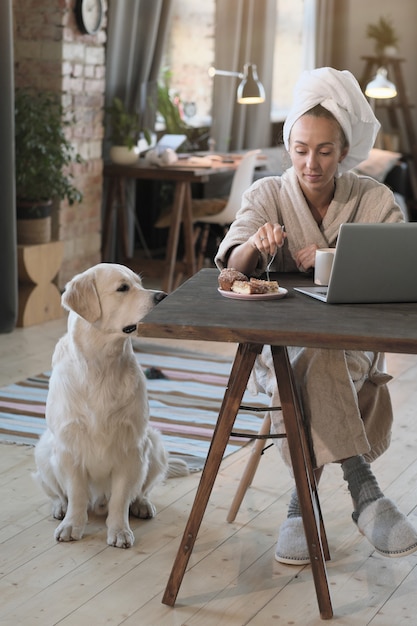 Jeune femme en peignoir assis à la table devant un ordinateur portable et boire du café avec un chien assis près d'elle à la maison
