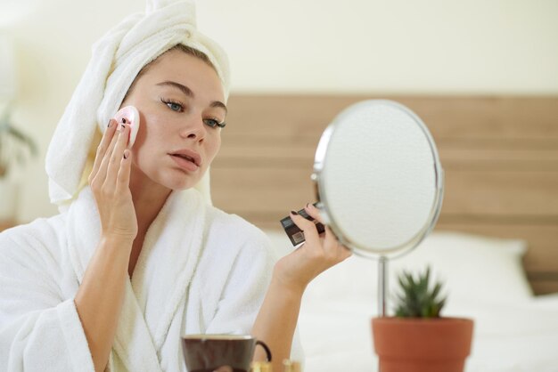 Jeune femme en peignoir regardant un miroir sur sa vanité et appliquant un fond de teint en poudre