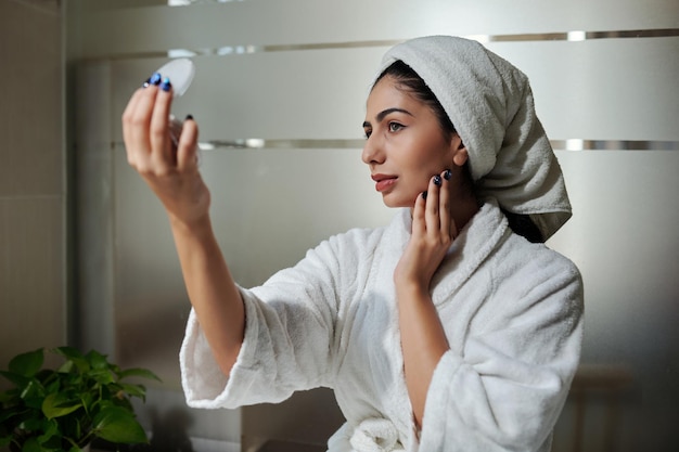 Jeune femme en peignoir et serviette sur la tête regardant un miroir compact