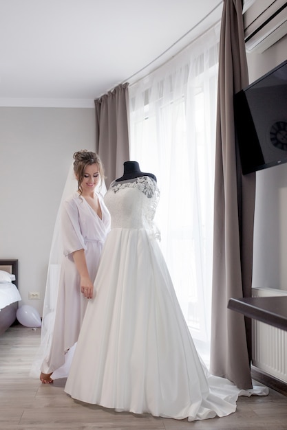 Jeune femme en peignoir de soie touchant sa robe de mariée et souriant tout en se tenant près de la fenêtre. Jour de mariage. Beauté, mode. Jour de mariage le plus heureux. Apprécie chaque moment.