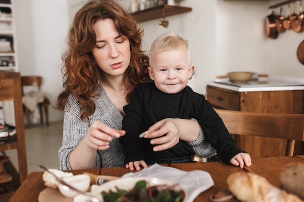 Jeune femme pensive aux cheveux rouges en pull tricoté assis à la table avec son petit fils souriant qui heureusement