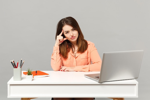 Jeune femme pensive dans des vêtements décontractés pastel mettant la main sur la tête, s'asseoir au bureau blanc avec ordinateur portable pc isolé sur fond gris. Concept de style de vie de carrière d'affaires de réalisation. Maquette de l'espace de copie.