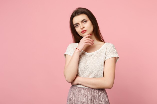 Jeune femme pensive dans des vêtements légers décontractés posant isolé sur fond rose, portrait en studio. Concept de mode de vie des émotions sincères des gens. Maquette de l'espace de copie. Regarder la caméra mettre la main sur le menton.