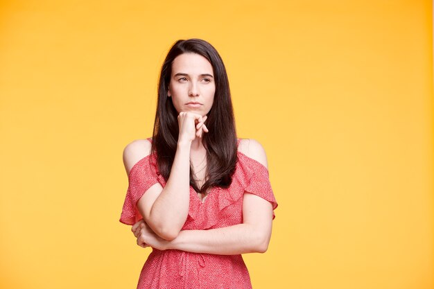 Jeune femme pensive ou mécontentement en robe élégante rouge en gardant une main par le menton tout en posant sur un mur jaune