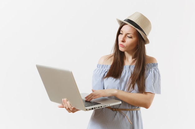 Jeune femme pensive en robe bleue, chapeau travaillant sur ordinateur portable moderne isolé sur fond blanc. Personnes, affaires indépendantes, style de vie, concept d'achat en ligne. Bureau mobile. Publicité.