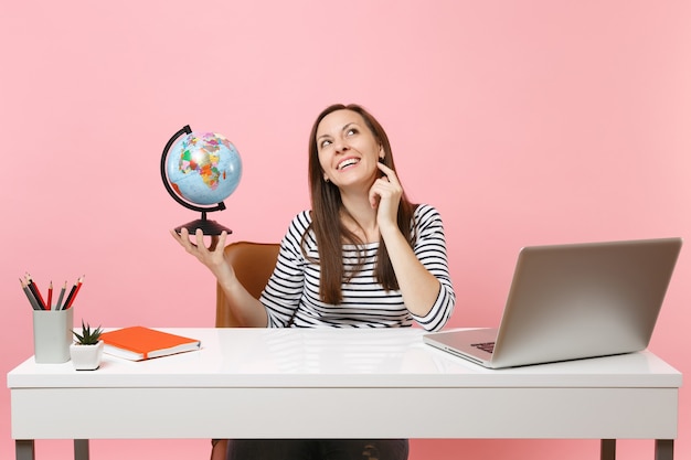 Jeune femme pensive tenant un globe, rêvant de planifier des vacances assis, travaille au bureau avec un ordinateur portable contemporain isolé sur fond rose pastel. Concept de carrière d'entreprise de réalisation. Espace de copie.