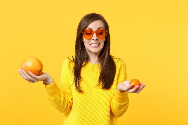 Jeune femme perplexe dans des lunettes de coeur tenant dans les mains des mandarines mûres fraîches, des fruits orange isolés sur fond jaune en studio. Mode de vie vivant des gens, concept de vacances relaxantes. Maquette de l'espace de copie.