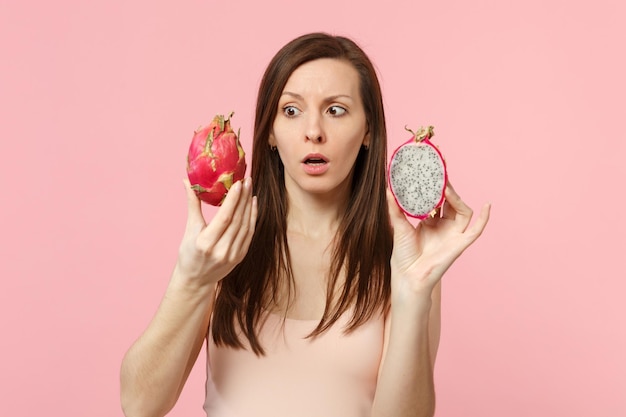 Jeune femme perplexe tenant à la main des moitiés de pitahaya mûre fraîche, fruit du dragon isolé sur fond de mur pastel rose en studio. Les gens vivent un style de vie vivant et se détendent. Maquette de l'espace de copie.