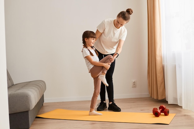 Jeune femme avec une petite fille portant des vêtements de sport faisant des exercices de sport à la maison enfant debout avec une jambe levée cuisses entraînement échauffement muscles d'entraînement enfant s'entraînant avec un entraîneur