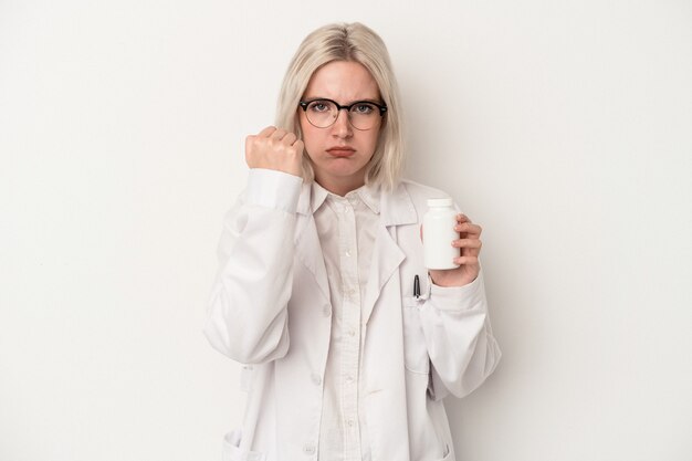 Jeune femme pharmacienne tenant des pilules isolées sur fond blanc montrant le poing à la caméra, expression faciale agressive.