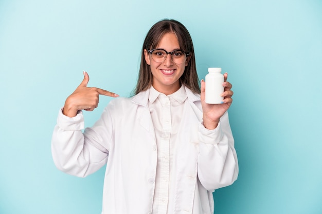 Jeune femme pharmacienne tenant des pilules isolées sur fond bleu personne pointant à la main vers un espace de copie de chemise, fière et confiante