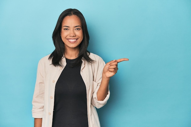 Jeune femme philippine sur studio bleu souriant joyeusement pointant avec l'index loin