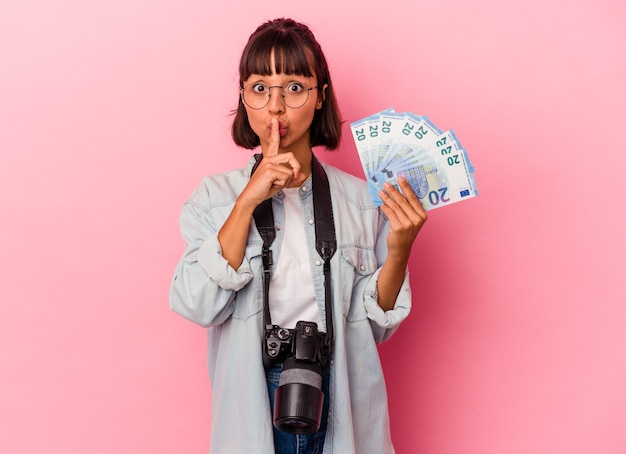 Jeune femme photographe métisse tenant des factures isolées sur fond rose gardant un secret ou demandant le silence.