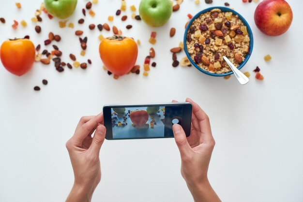 Jeune femme photographiant son petit déjeuner. Fille mangeant des céréales de petit déjeuner avec des noix, des graines de citrouille, de l'avoine et dans un bol avec des fruits. Fille tenant un téléphone intelligent. Collation saine ou petit-déjeuner le matin.