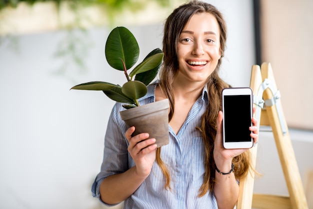 Jeune femme plantant une maison avec de la verdure debout avec un téléphone et un pot de fleurs sur l'échelle