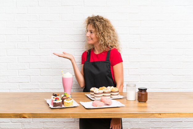 Jeune femme avec plein de mini gâteaux différents sur une table, étendant les mains sur le côté pour inviter à venir