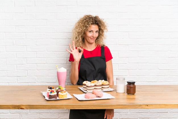 Jeune femme avec plein de mini gâteaux différents sur une table montrant un signe ok avec les doigts