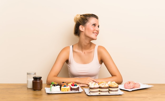 Jeune femme avec plein de mini gâteaux différents sur une table en riant et levant les yeux
