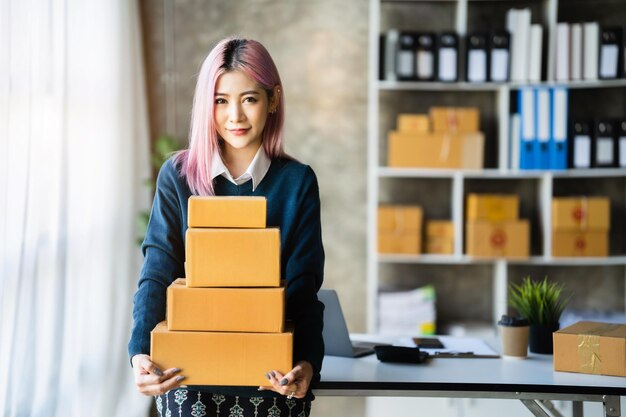 Jeune femme PME asiatique tenant des colis et regardant la caméra debout parmi plusieurs boîtes dans un bureau moderne