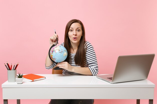 Jeune femme pointant sur un globe avec un crayon, planifiant des vacances tout en s'asseyant et travaillant au bureau blanc avec un ordinateur portable contemporain