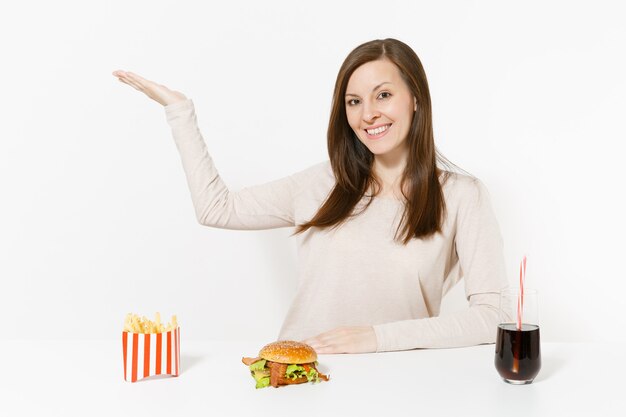 Jeune femme pointant la main de côté sur l'espace de copie à table avec hamburger, frites, cola en bouteille de verre isolé sur fond blanc. Une bonne nutrition ou une restauration rapide classique américaine. Espace publicitaire.