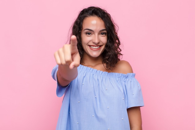 Jeune femme pointant avec un sourire satisfait, confiant et amical, vous choisissant sur le mur rose