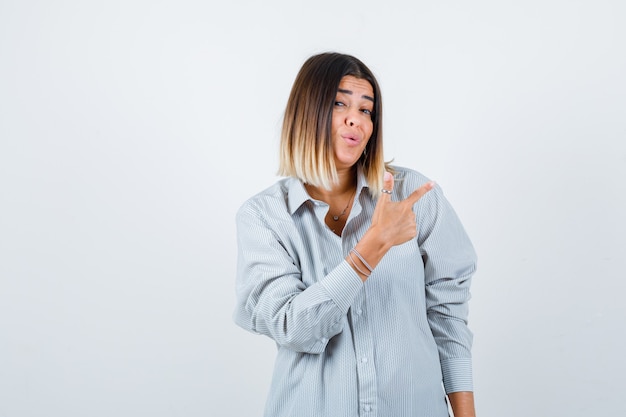 Jeune femme pointant vers le coin supérieur droit en chemise surdimensionnée et l'air confiant, vue de face.