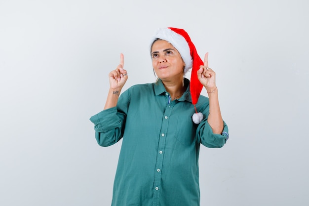 Jeune femme pointant vers le haut en chemise, bonnet de Noel et l'air confiant, vue de face.