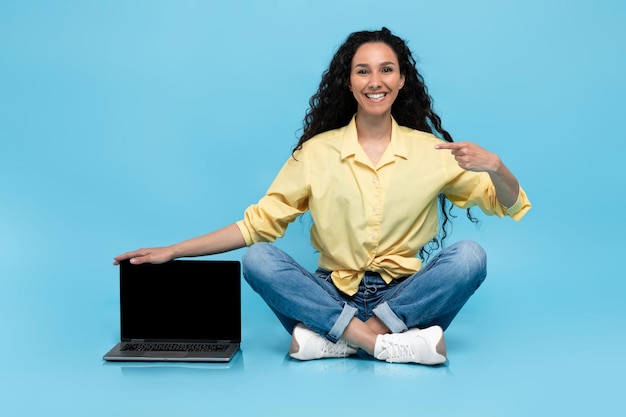 Jeune femme pointant vers un ordinateur portable avec écran vide offrant une maquette pour site Web sur studio bleu