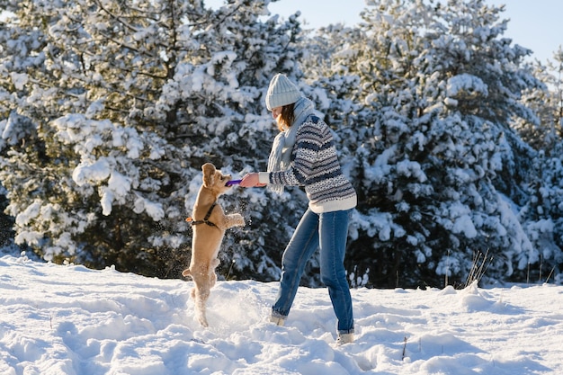 Une jeune femme portant un chandail et une écharpe bonnet tricoté joue avec son chien dans la forêt d'hiver Journée ensoleillée Le chien saute pour un jouet