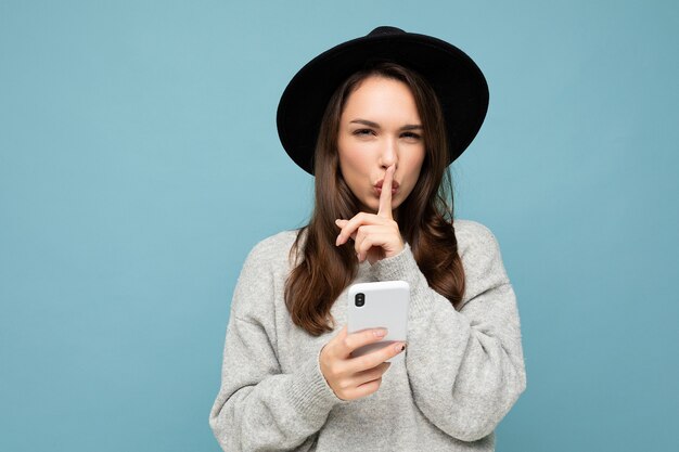 Jeune femme portant un chapeau noir et un pull gris tenant un smartphone regardant la caméra montrant chut